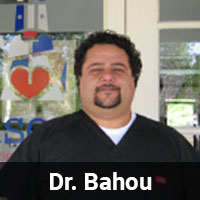 A man with curly hair and a beard stands in front of a glass door, wearing a black medical scrub top. The name tag reads "Dr. Bahou" at the bottom of the image, indicating he's a veterinarian. The background includes a partial logo with a heart and other colorful shapes.