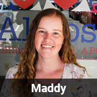 A person with long, wavy hair is smiling at the camera. They are standing in front of a glass door with text and some images partially visible in the background. The name "Maddy, Vet" is displayed in bold white letters at the bottom.