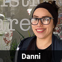 A person with glasses, a headband, and a black jacket is smiling in front of a window with text and some visible plants inside. The name "Danni" is displayed at the bottom of the image, hinting that this could be our friendly local vet.