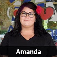 A person with long dark hair, wearing glasses and a red flower in their hair, is smiling and standing in front of a backdrop with colorful heart and smiley face designs. The name "Amanda" is displayed in a black bar at the bottom of the image, showcasing her pride as a dedicated veterinarian.