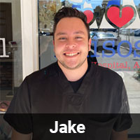 A man named Jake is smiling broadly while standing in front of a glass door. He is wearing black scrubs, and the name "Jake" is displayed at the bottom of the image. In the background, part of a sign with red hearts and some text is visible on the door, hinting that Jake might be a caring veterinarian.