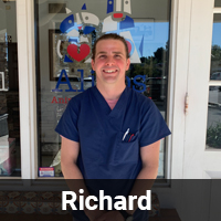 A veterinarian in blue scrubs stands in front of a glass door with the name "Richard" displayed at the bottom of the image. The door features a logo with a heart and paw print, suggesting this is a veterinary clinic or animal care facility.